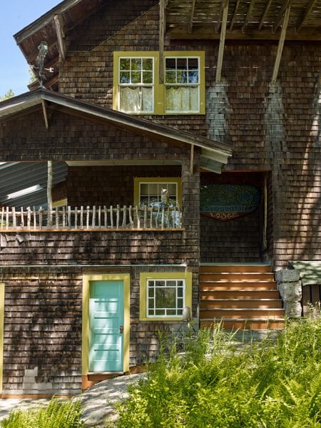 A view of the side approach with focus on the natural Birch logs, used as a railing on the upper deck, and vibrant trim around the doors and windows.