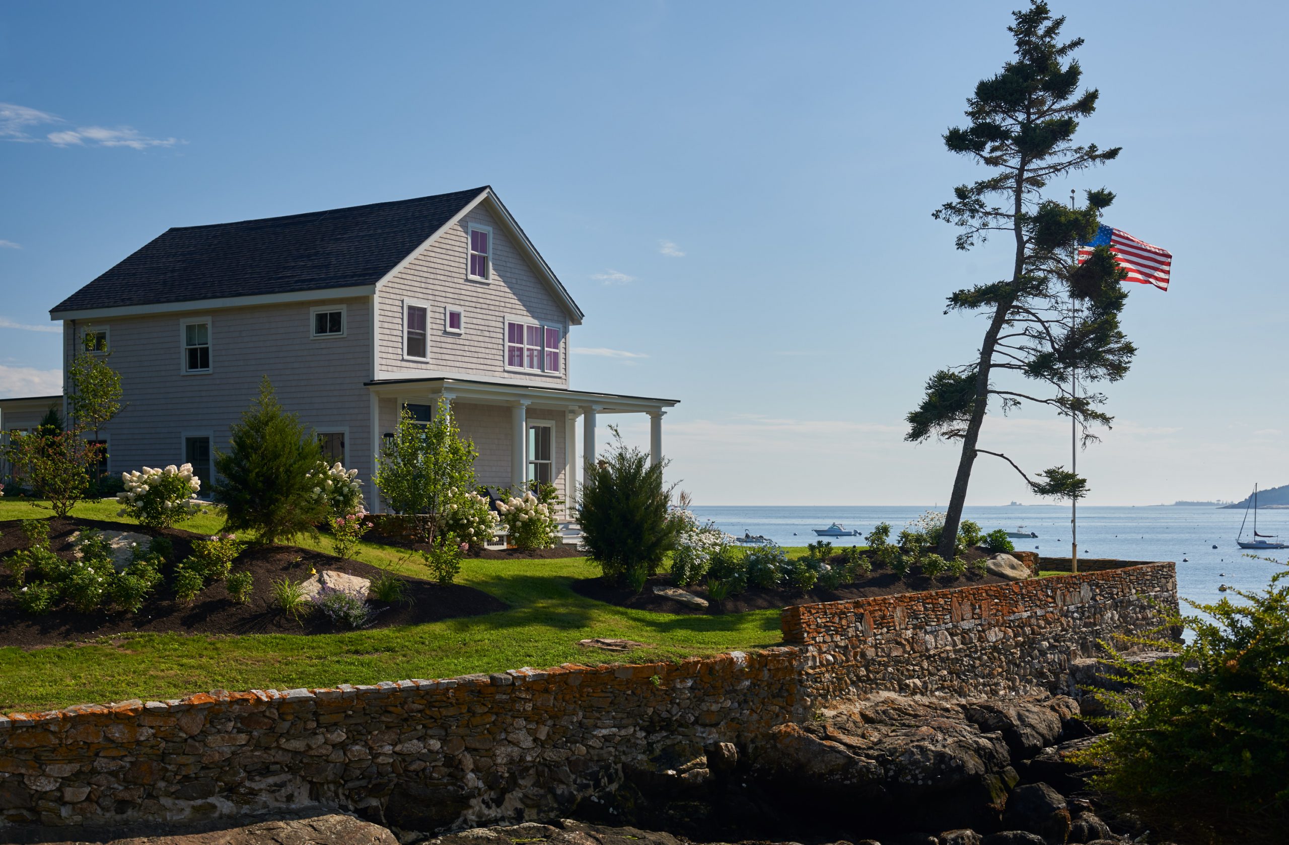A stunning Twin Cove home with spacious lawn, wood side detailing, a flagpole, and a stone wall that wraps along the coastline.
